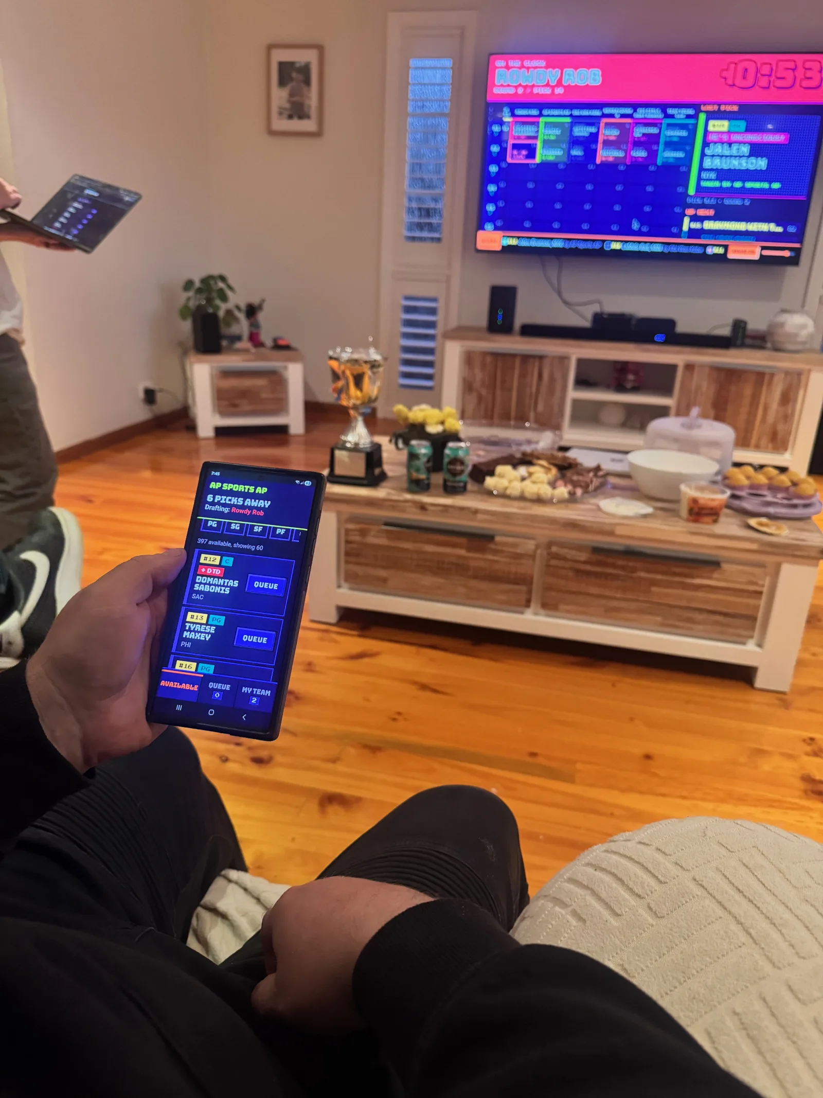 Someone on the couch checking their queue on a phone, six picks away, with the board on the TV behind